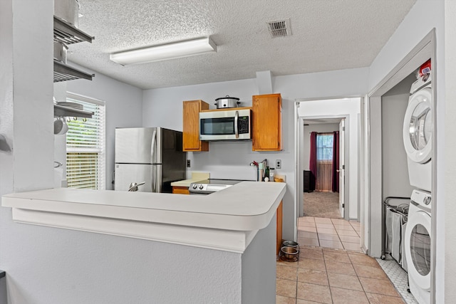 kitchen featuring kitchen peninsula, appliances with stainless steel finishes, a textured ceiling, light tile patterned floors, and stacked washer and clothes dryer