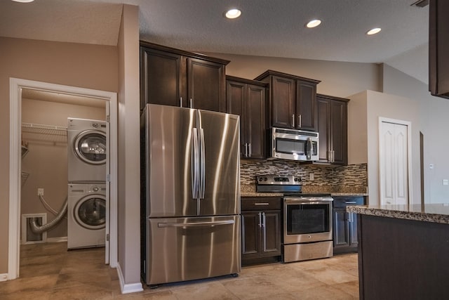 kitchen with stacked washing maching and dryer, dark stone countertops, appliances with stainless steel finishes, lofted ceiling, and backsplash