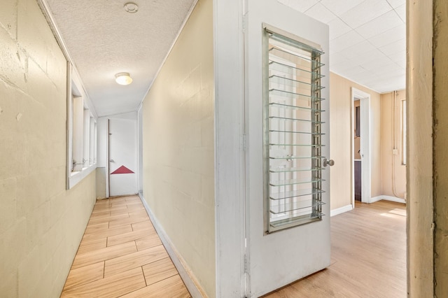 hall with a textured ceiling, light wood finished floors, concrete block wall, and baseboards