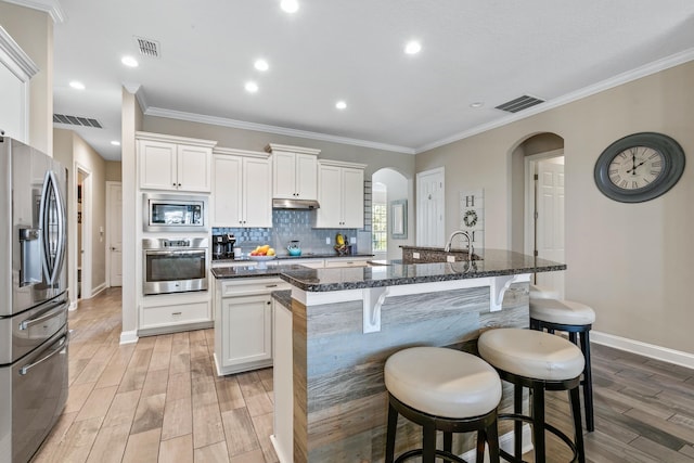 kitchen with white cabinetry, dark stone countertops, decorative backsplash, stainless steel appliances, and a center island with sink