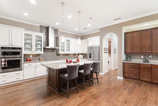 kitchen featuring a breakfast bar, hanging light fixtures, stainless steel appliances, a kitchen island with sink, and wall chimney range hood