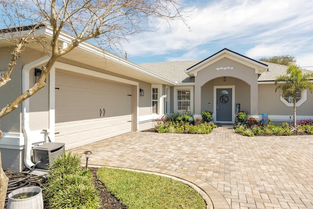 ranch-style home featuring a garage, central AC, roof with shingles, decorative driveway, and stucco siding