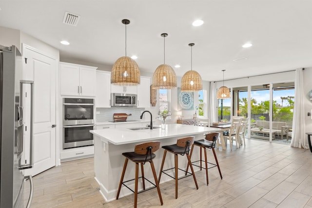 kitchen featuring sink, appliances with stainless steel finishes, white cabinetry, hanging light fixtures, and a center island with sink