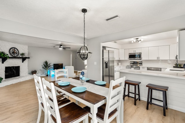 dining space featuring visible vents, a stone fireplace, light wood finished floors, and ceiling fan with notable chandelier