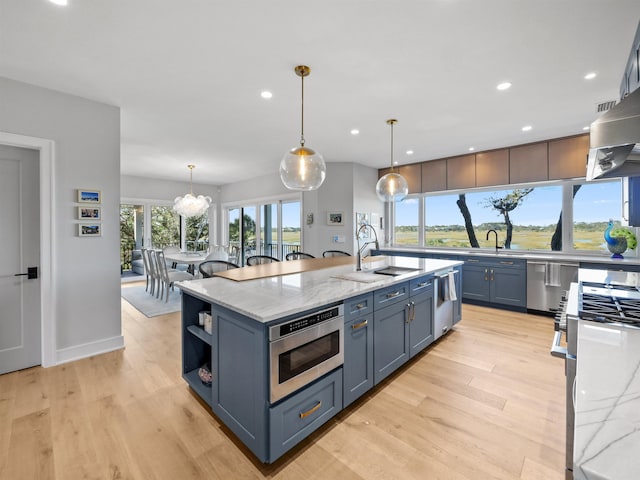 kitchen with light stone countertops, light wood-type flooring, decorative light fixtures, stainless steel appliances, and a center island with sink