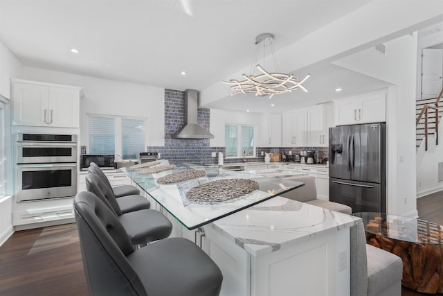 kitchen with refrigerator with ice dispenser, a kitchen island, double oven, an inviting chandelier, and wall chimney exhaust hood