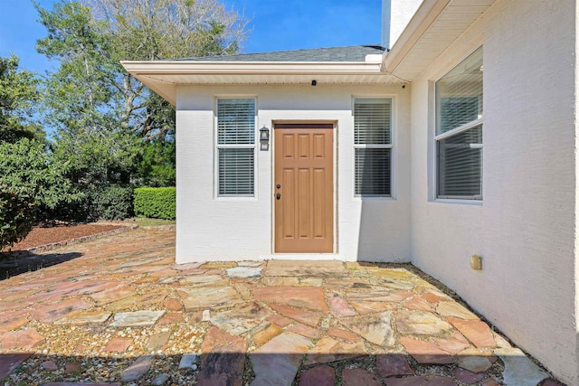 view of exterior entry featuring a shingled roof, a patio area, and stucco siding