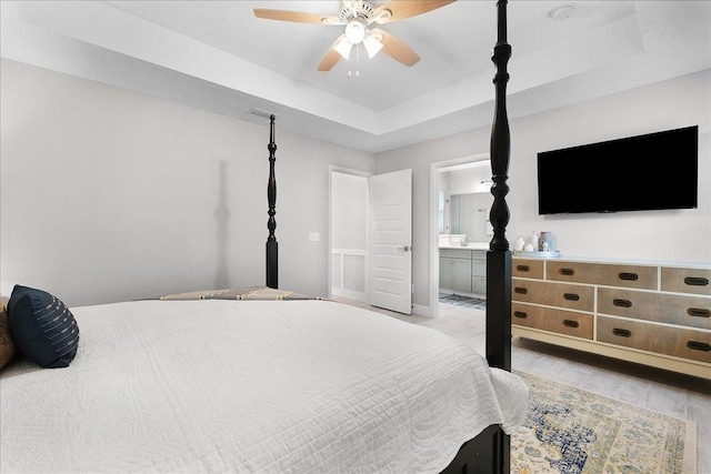 bedroom featuring ensuite bath, light wood-style flooring, a tray ceiling, and a ceiling fan