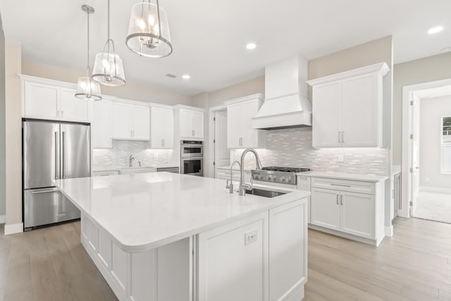 kitchen featuring custom exhaust hood, decorative light fixtures, an island with sink, stainless steel appliances, and white cabinets