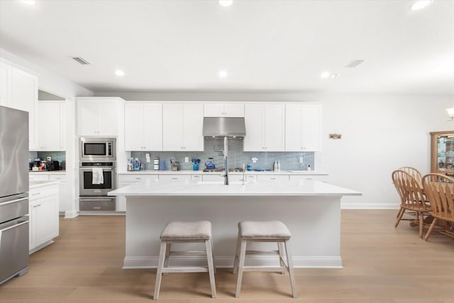 kitchen featuring appliances with stainless steel finishes, a kitchen island with sink, backsplash, white cabinetry, and light wood-type flooring