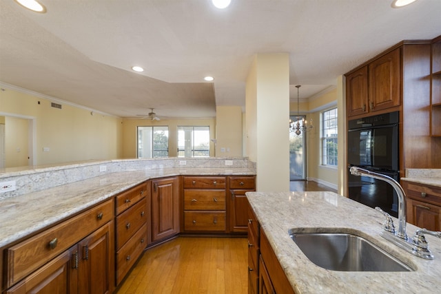 kitchen with light stone countertops, ceiling fan with notable chandelier, double oven, sink, and light hardwood / wood-style flooring