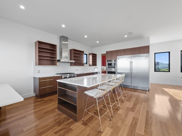 kitchen with a center island with sink, a kitchen breakfast bar, wall chimney range hood, appliances with stainless steel finishes, and light hardwood / wood-style floors