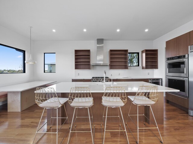 kitchen featuring a kitchen bar, wall chimney exhaust hood, double oven, wood-type flooring, and an island with sink