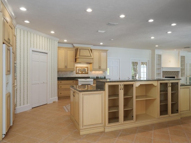 kitchen with light brown cabinetry, backsplash, custom range hood, white appliances, and a kitchen island with sink