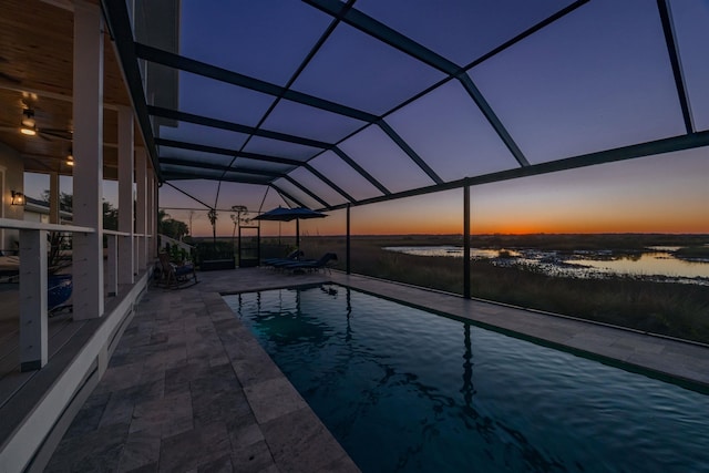 pool at dusk featuring a water view, glass enclosure, and a patio area