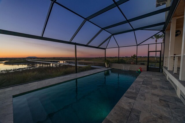 pool at dusk featuring glass enclosure, a patio area, and a water view