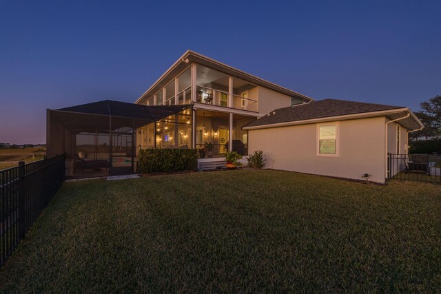 back house at dusk featuring a balcony, glass enclosure, and a lawn