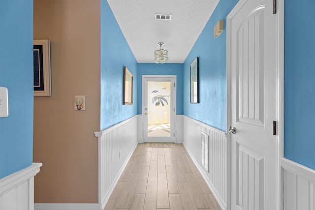 corridor with light wood-type flooring, a wainscoted wall, visible vents, and a textured ceiling