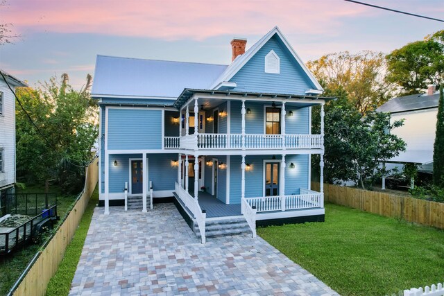 view of front of home featuring a balcony, covered porch, fence, a lawn, and a chimney