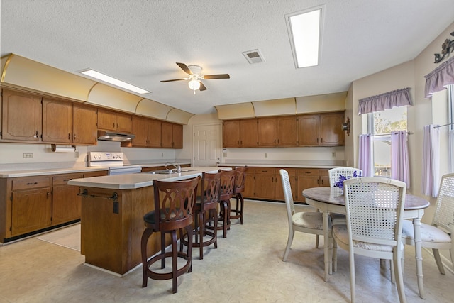 kitchen with ceiling fan, a kitchen breakfast bar, white range with electric stovetop, a textured ceiling, and a kitchen island with sink