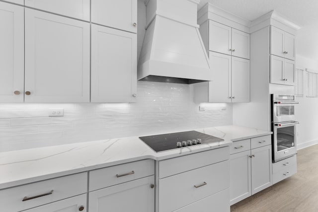 kitchen with custom range hood, white cabinetry, double oven, black electric cooktop, and backsplash