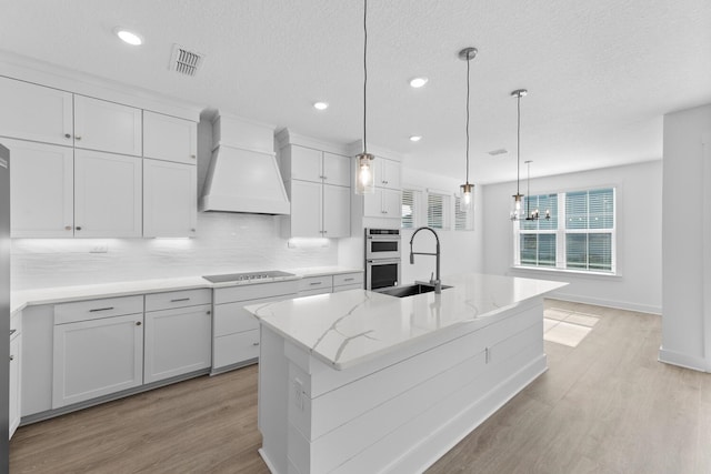 kitchen featuring premium range hood, light wood-style flooring, a sink, double oven, and decorative backsplash