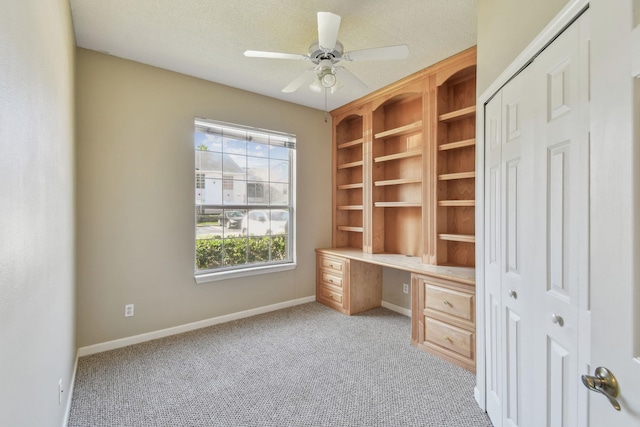 unfurnished office with baseboards, ceiling fan, built in desk, a textured ceiling, and light colored carpet