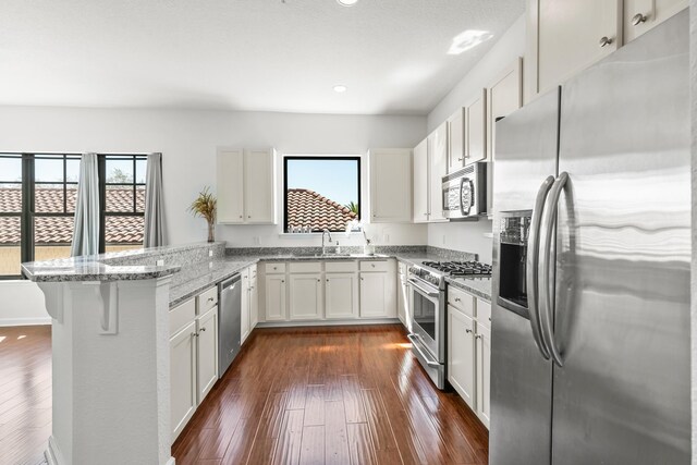 kitchen with kitchen peninsula, appliances with stainless steel finishes, light stone countertops, dark hardwood / wood-style floors, and white cabinetry