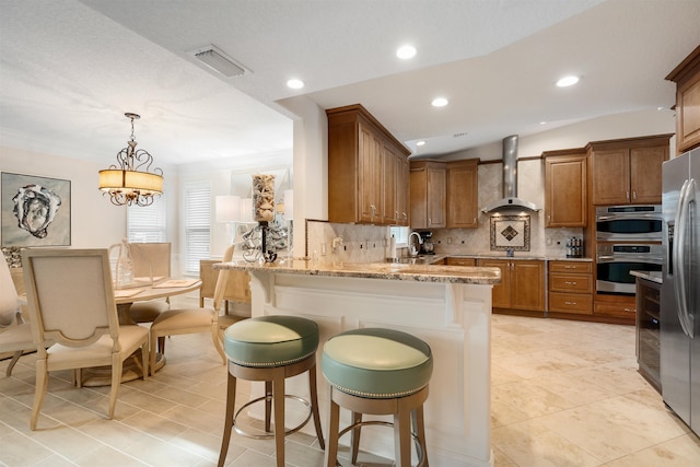 kitchen with appliances with stainless steel finishes, a kitchen breakfast bar, light stone counters, kitchen peninsula, and wall chimney exhaust hood