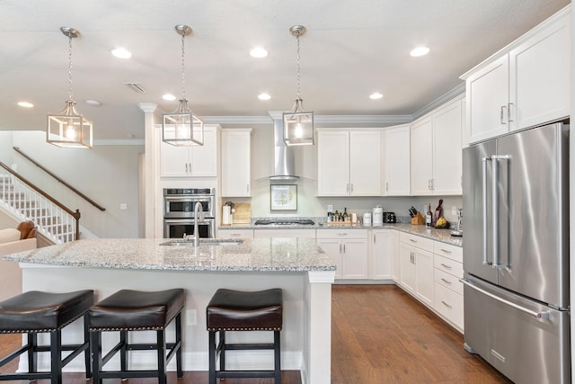 kitchen featuring white cabinets, wall chimney exhaust hood, decorative light fixtures, stainless steel appliances, and an island with sink