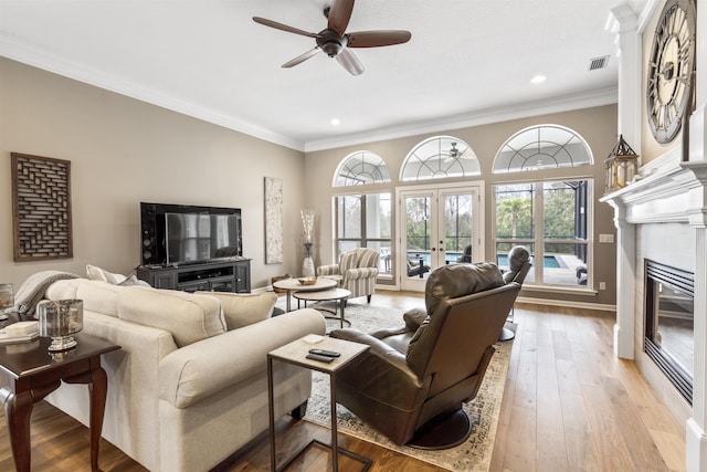 living area featuring light wood finished floors, a ceiling fan, a tile fireplace, crown molding, and french doors