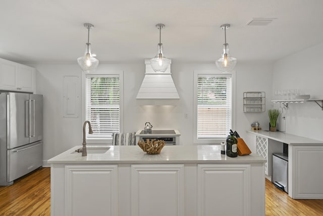 kitchen with sink, high end fridge, an island with sink, white cabinets, and decorative light fixtures