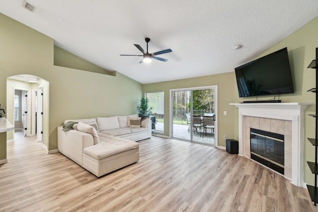 living room featuring arched walkways, light wood finished floors, lofted ceiling, visible vents, and a tiled fireplace