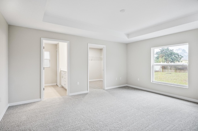 unfurnished bedroom with a spacious closet, light colored carpet, a tray ceiling, ensuite bath, and a closet