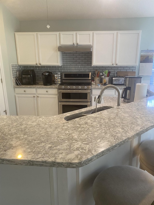 kitchen with under cabinet range hood, a sink, tasteful backsplash, white cabinetry, and range with two ovens