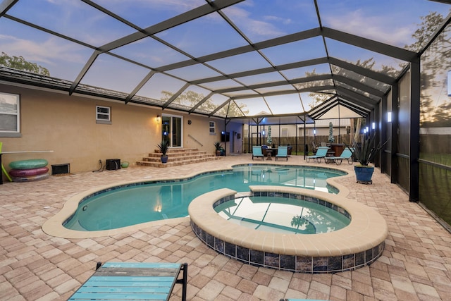 pool at dusk featuring glass enclosure, an in ground hot tub, and a patio area