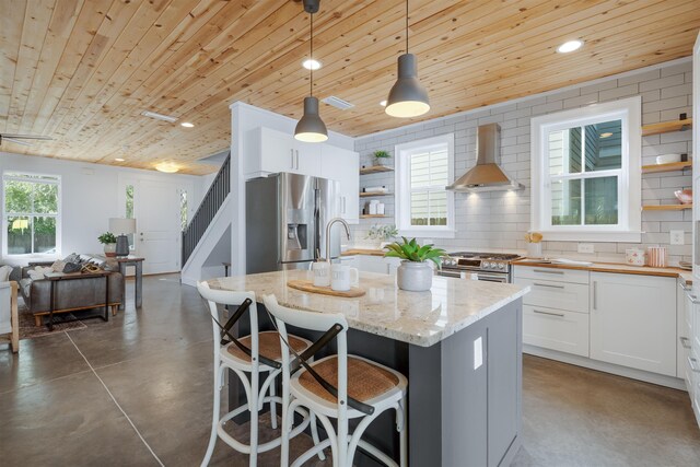 kitchen featuring light stone counters, wall chimney exhaust hood, white cabinets, hanging light fixtures, and an island with sink