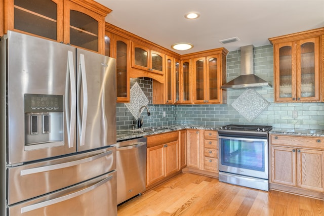 kitchen with a sink, stainless steel appliances, light wood-style floors, wall chimney range hood, and stone counters