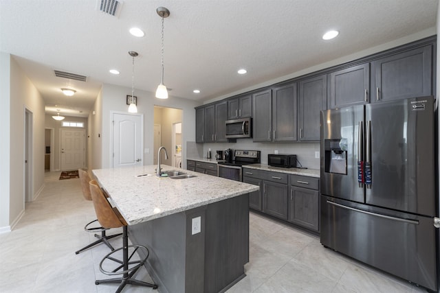 kitchen with sink, stainless steel appliances, light stone counters, an island with sink, and decorative light fixtures