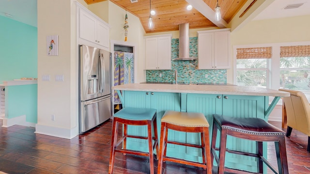 kitchen featuring wall chimney exhaust hood, stainless steel refrigerator with ice dispenser, a kitchen bar, white cabinetry, and wooden ceiling