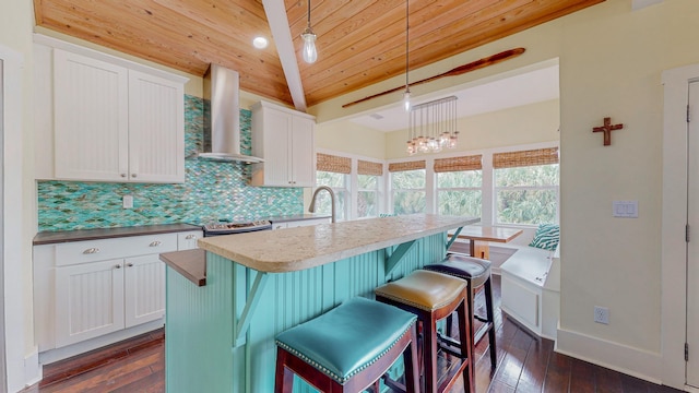 kitchen featuring decorative light fixtures, white cabinetry, wooden ceiling, wall chimney range hood, and a center island with sink