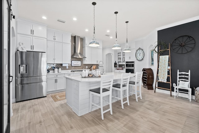 kitchen featuring appliances with stainless steel finishes, tasteful backsplash, white cabinetry, wall chimney range hood, and a center island with sink