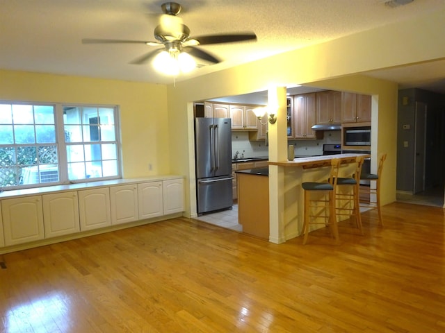 kitchen featuring under cabinet range hood, a ceiling fan, light wood-style floors, appliances with stainless steel finishes, and a kitchen bar