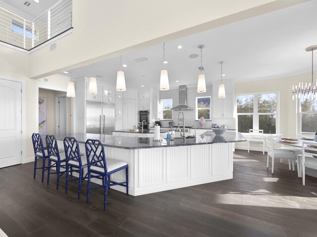 kitchen featuring built in fridge, wall chimney exhaust hood, a towering ceiling, decorative light fixtures, and white cabinetry