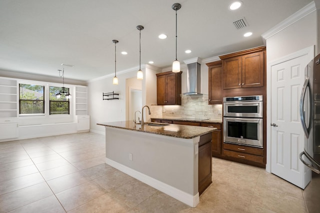 kitchen featuring wall chimney exhaust hood, stainless steel appliances, sink, decorative light fixtures, and an island with sink