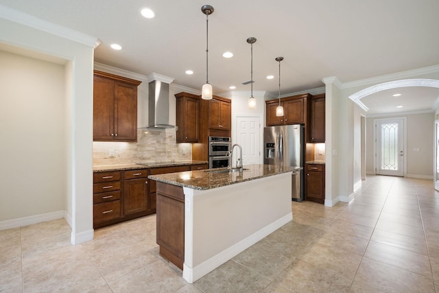 kitchen with wall chimney exhaust hood, hanging light fixtures, stainless steel appliances, dark stone countertops, and a center island with sink