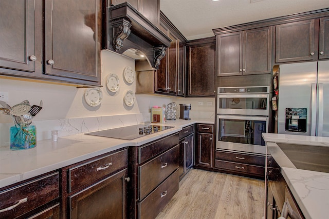 kitchen with dark brown cabinetry, stainless steel appliances, light stone counters, custom range hood, and light wood-type flooring