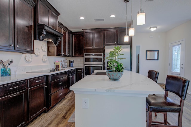 kitchen with light wood-type flooring, appliances with stainless steel finishes, decorative light fixtures, a kitchen island, and dark brown cabinetry