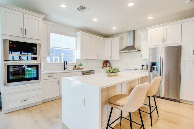 kitchen with wall chimney range hood, white cabinetry, stainless steel appliances, a kitchen island, and light wood-type flooring