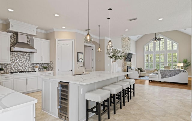 kitchen featuring wall chimney range hood, a large island with sink, beverage cooler, and decorative light fixtures